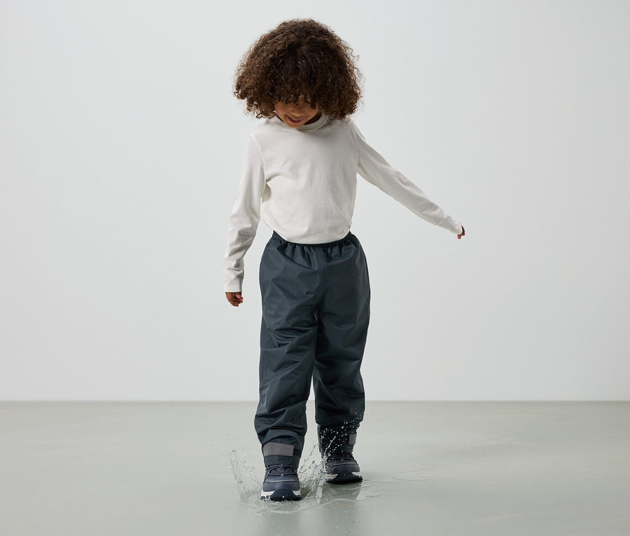G göndör hajú gyermek hosszú ujjú felsőben, szürke esőnadrágban és csizmában ugrál egy pocsolyába.