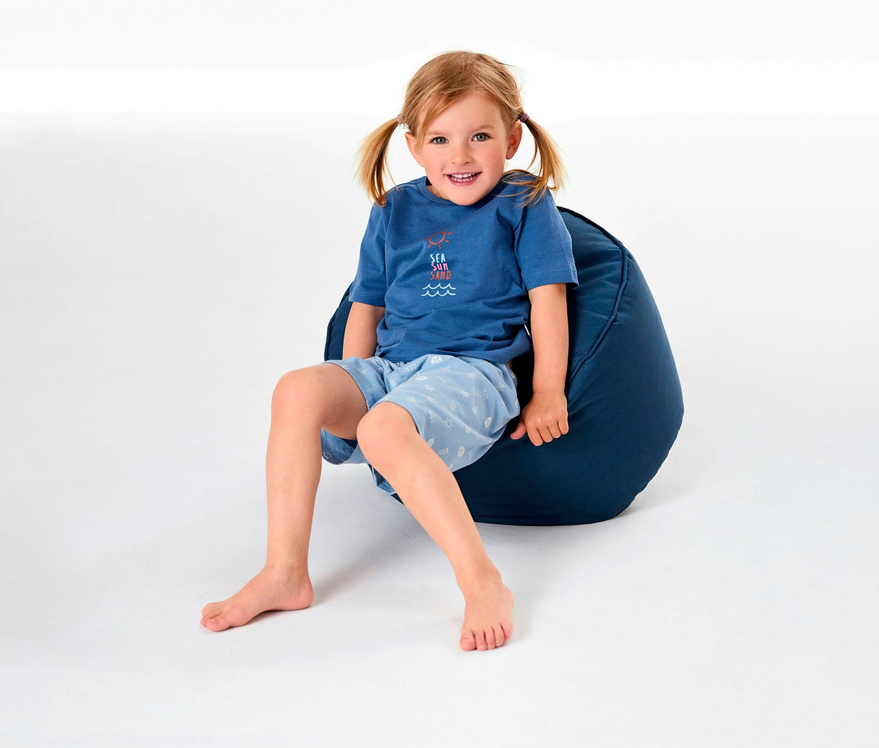 Egy kislány ül egy babzsákon, és SCHIESSER rövidnadrágos gyerekpizsamát visel.