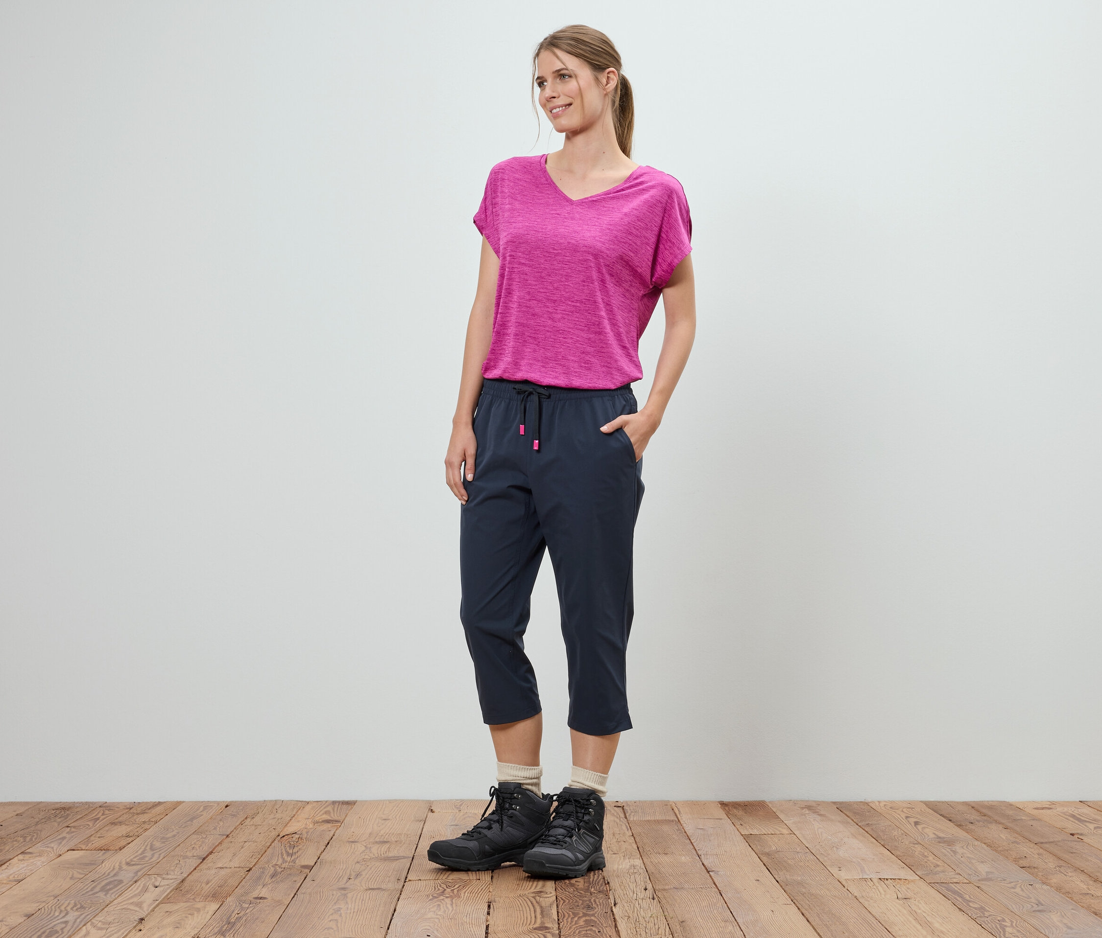 Egy nő áll a kezével a zsebében egy fehér fal előtt. Rózsaszín pólót, kék háromnegyedes nadrágot és fekete túrabakancsot visel.