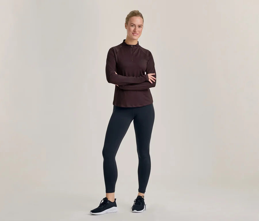 Egy mosolygó nő áll keresztbe tett karokkal, sötét, hosszú ujjú, félcipzáras felsőt, sötét leggingset és fekete-fehér sportcipőt visel.