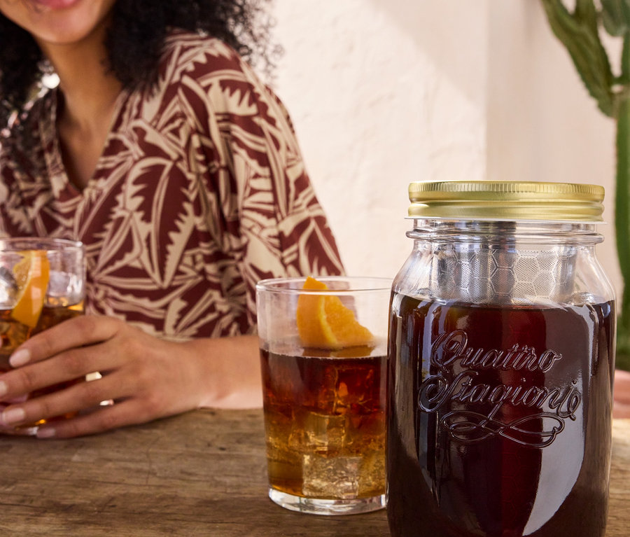 Nő itallal teli poharat tart a kezében a Cold Brew készítő »BrewJar« és a Női midiruha, levél, bézs/terrakotta mellett.