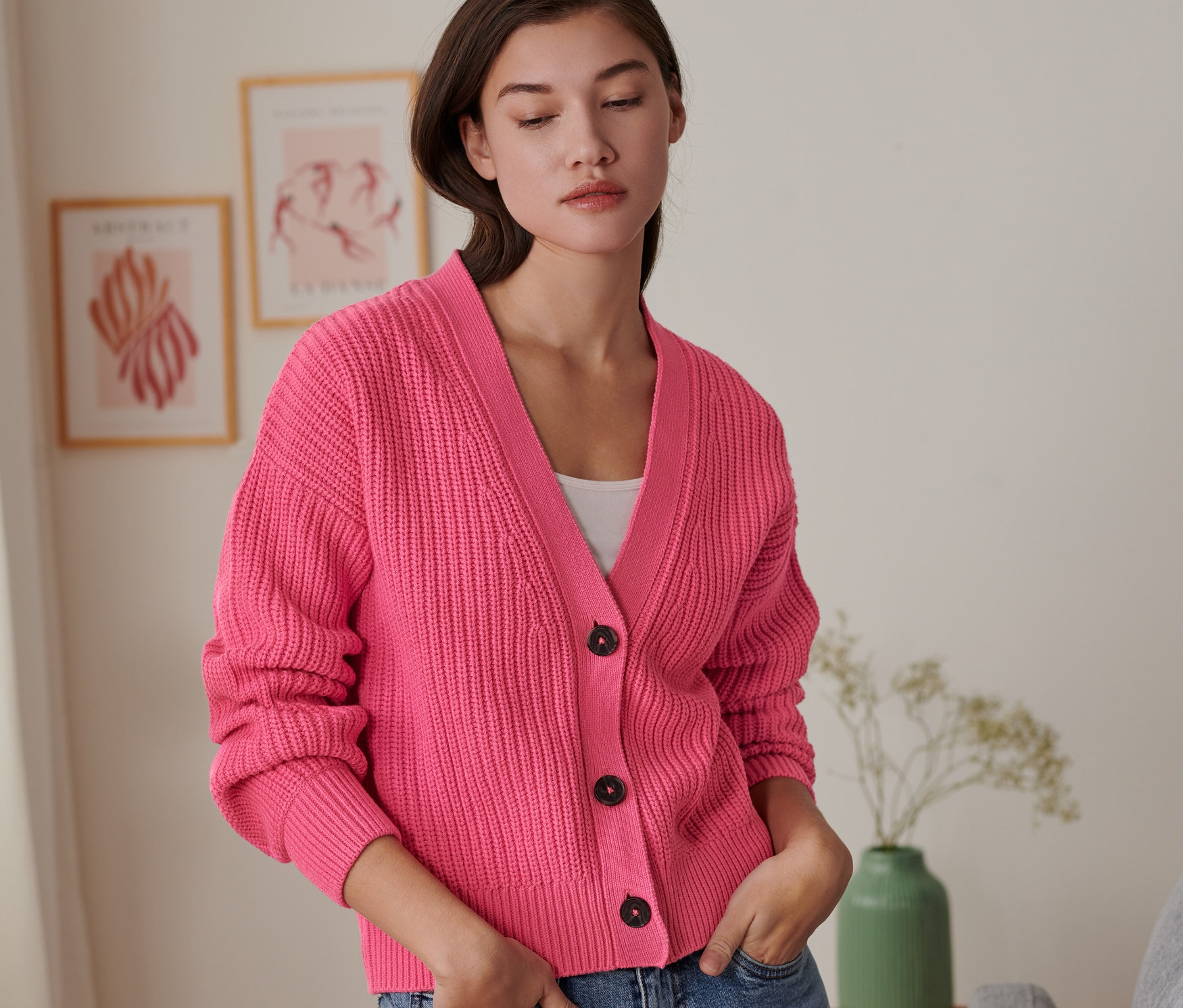 Egy nő farmernadrágjának zsebébe dugott kézzel rózsaszín kötött kardigánt visel.