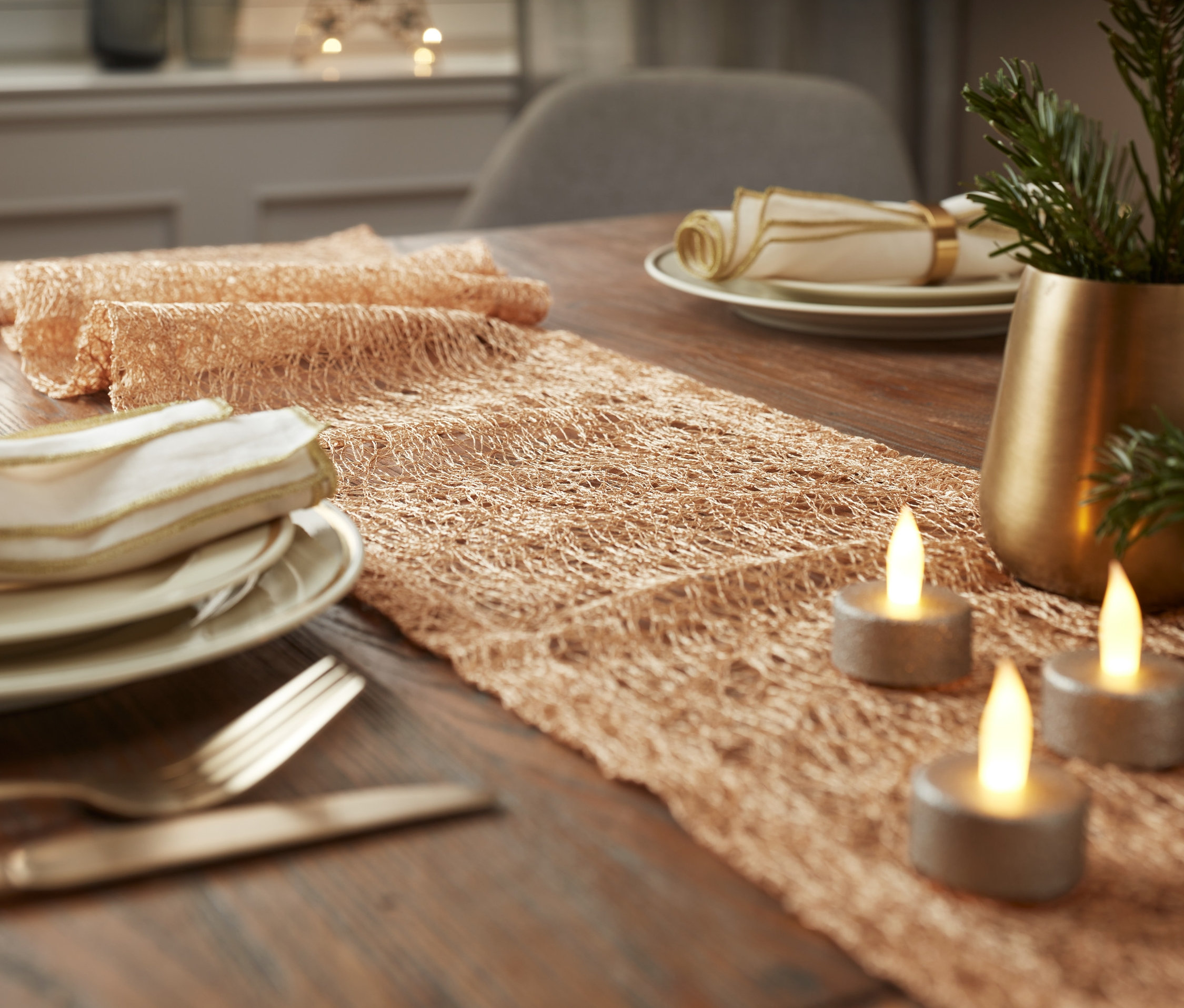 A fa asztalon egy bézs asztali futó, három égő gyertya és egy ágakkal teli váza látható.