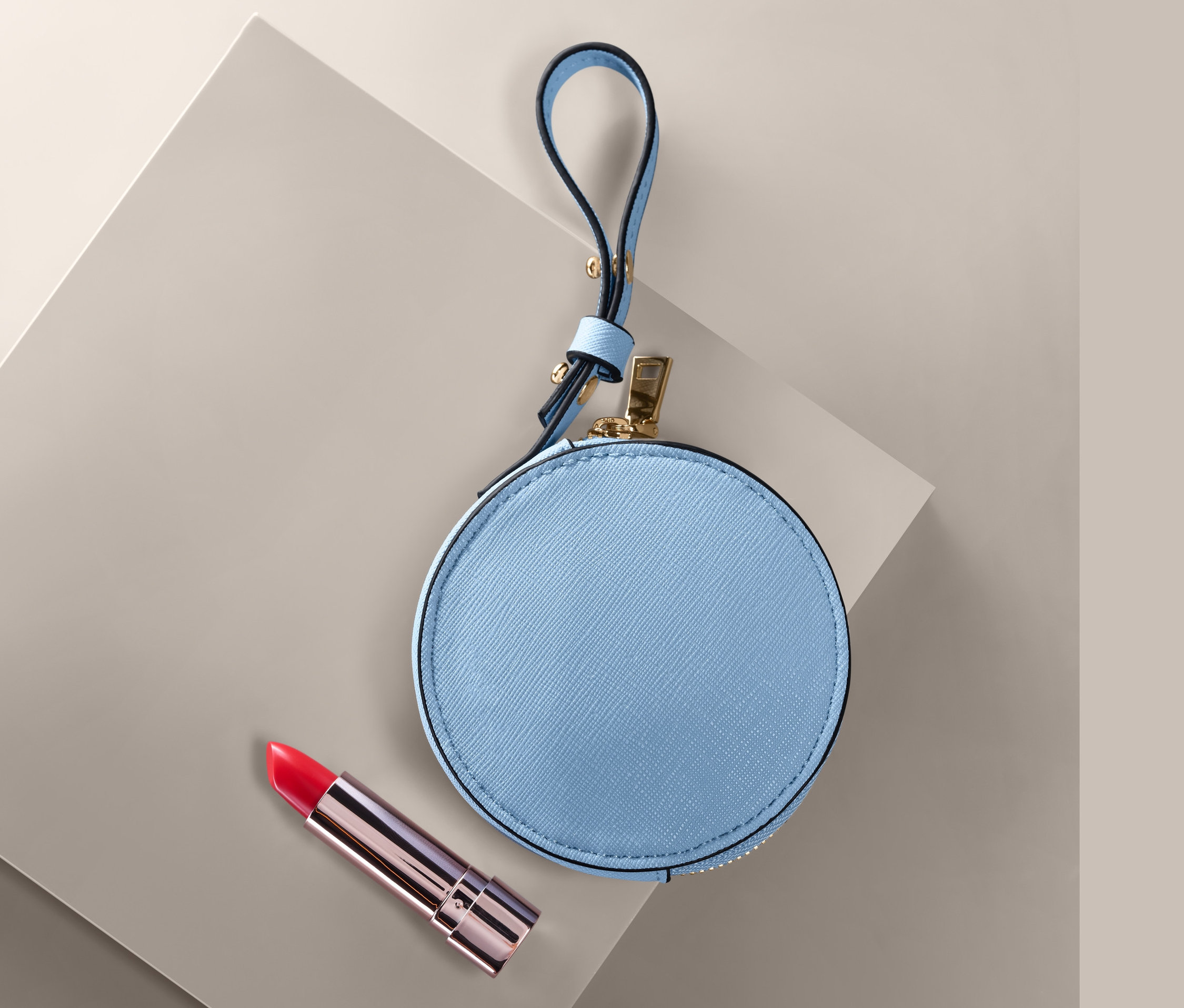 Egy kék, kerek női pénztárca és egy piros rúzs egy szürke felületen.