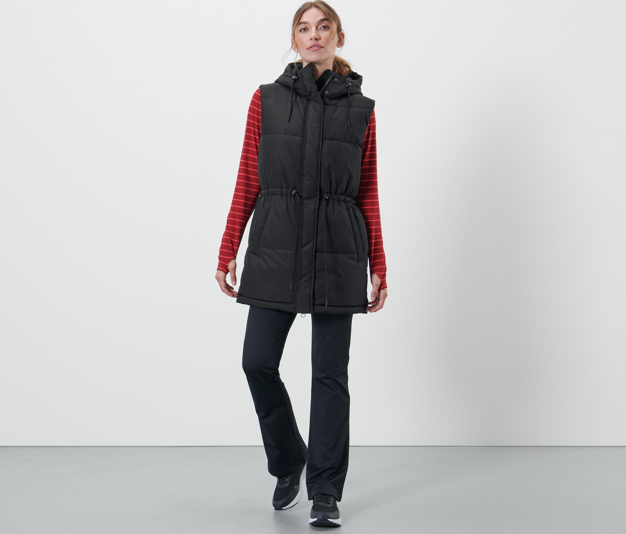 Nő visel egy antracit színű steppelt mellényt, egy piros csíkos hosszú ujjú inget és egy fekete nadrágot.