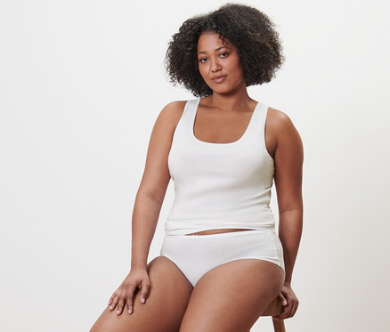 Nő ül egy széken fehér trikóban és alsóban. 5 db női trikó, fekete/fehér. 4 db női magas derekú alsó, fekete/fehér/testszínű.