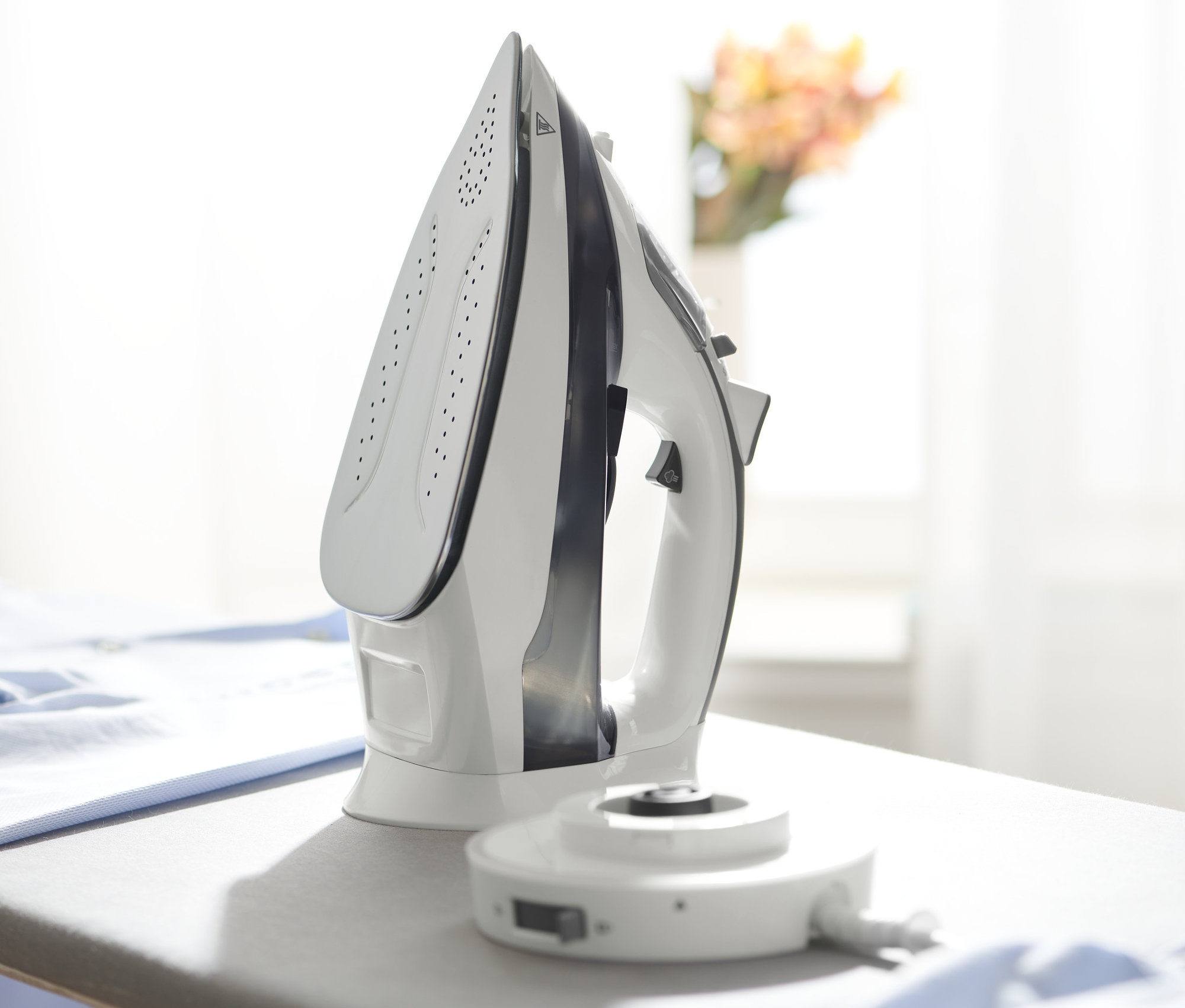 Egy fehér vezeték nélküli gőzölős vasaló áll egy szürke vasalódeszkán egy halom kék ruha mellett.