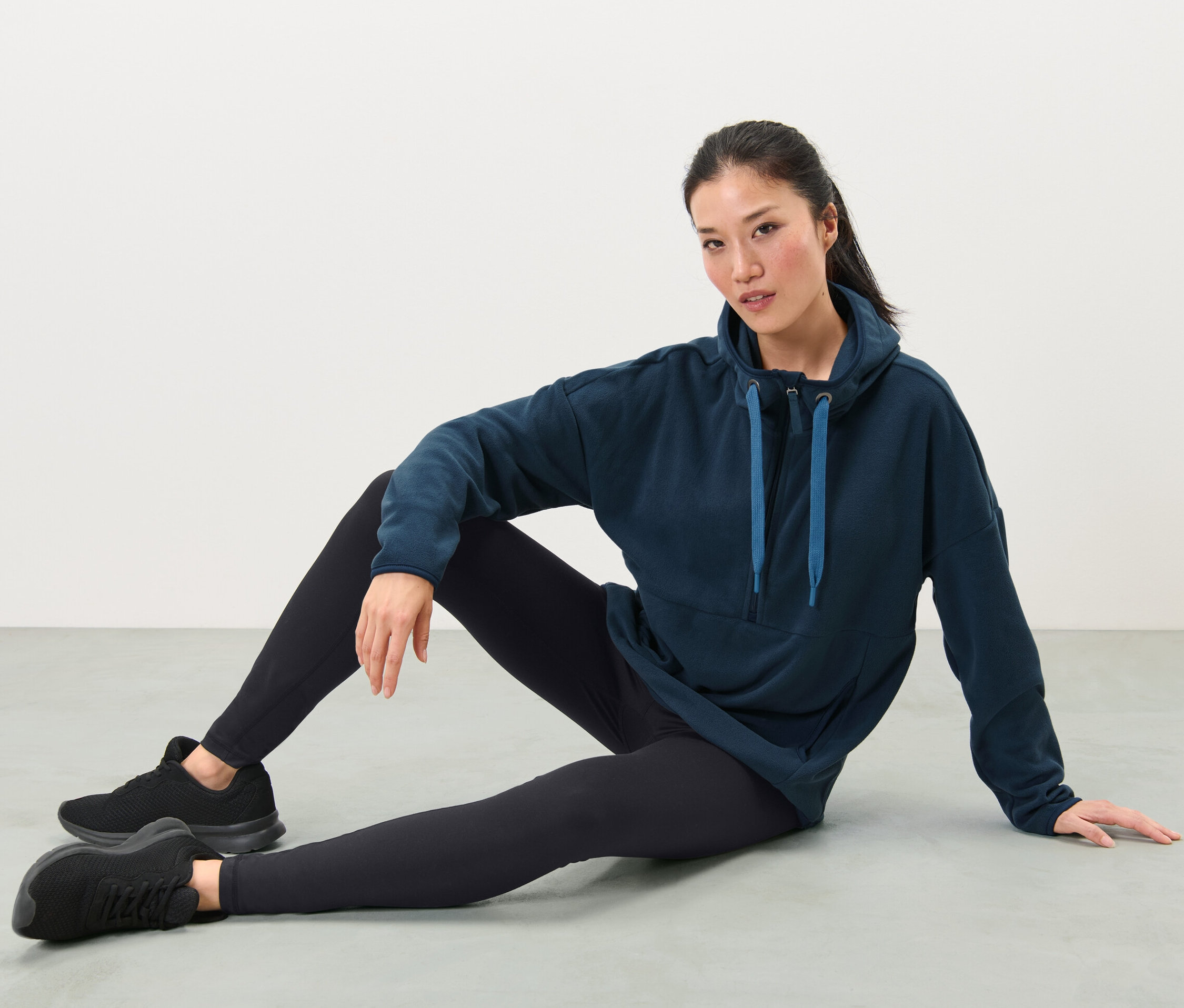 Nő ül a földön, sötétkék kapucnis mikropolár felsőt, fekete leggingset és edzőcipőt visel.