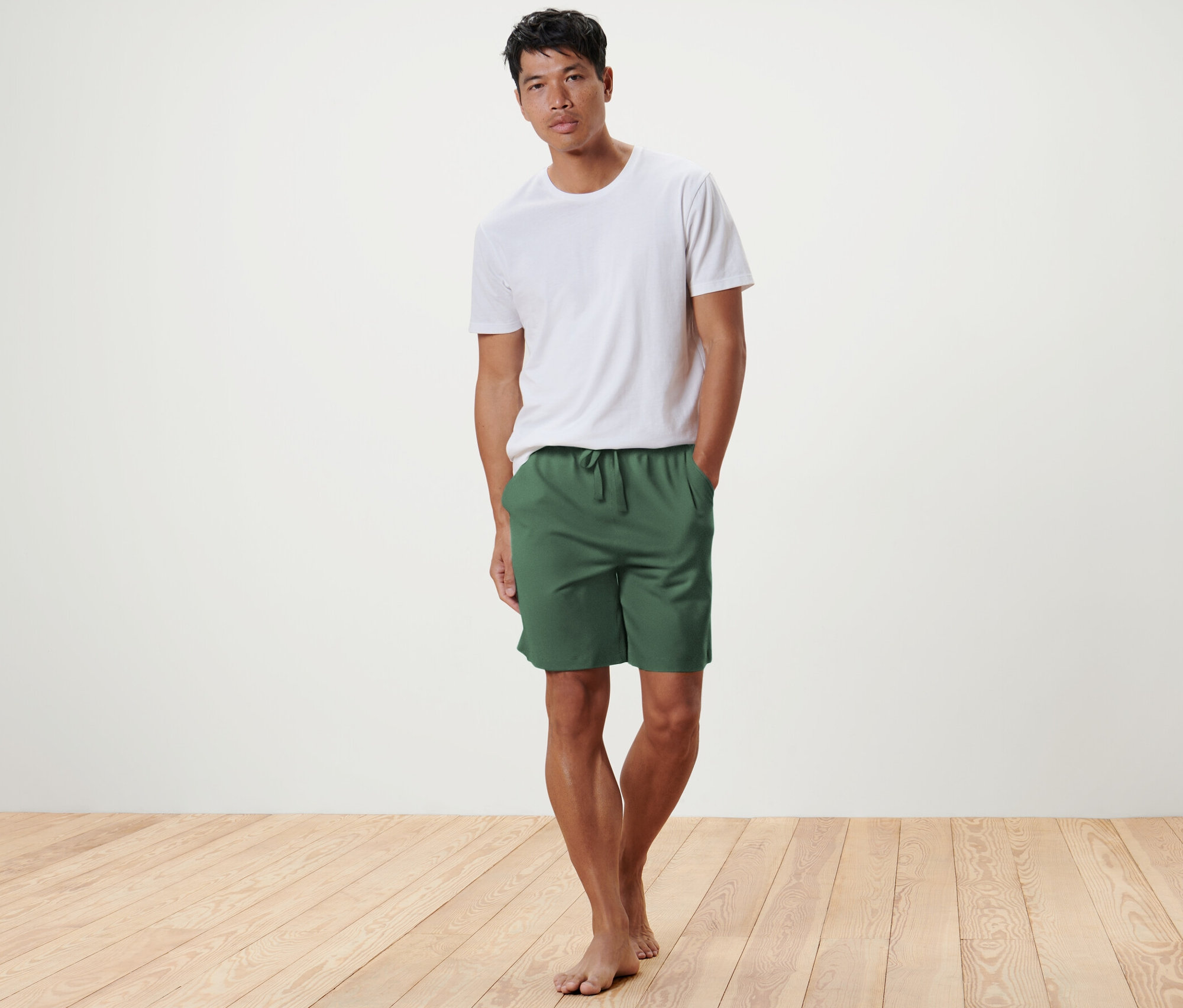 Férfi pózol a fapadlón olívazöld férfi jersey rövidnadrágban és fehér pólóban.