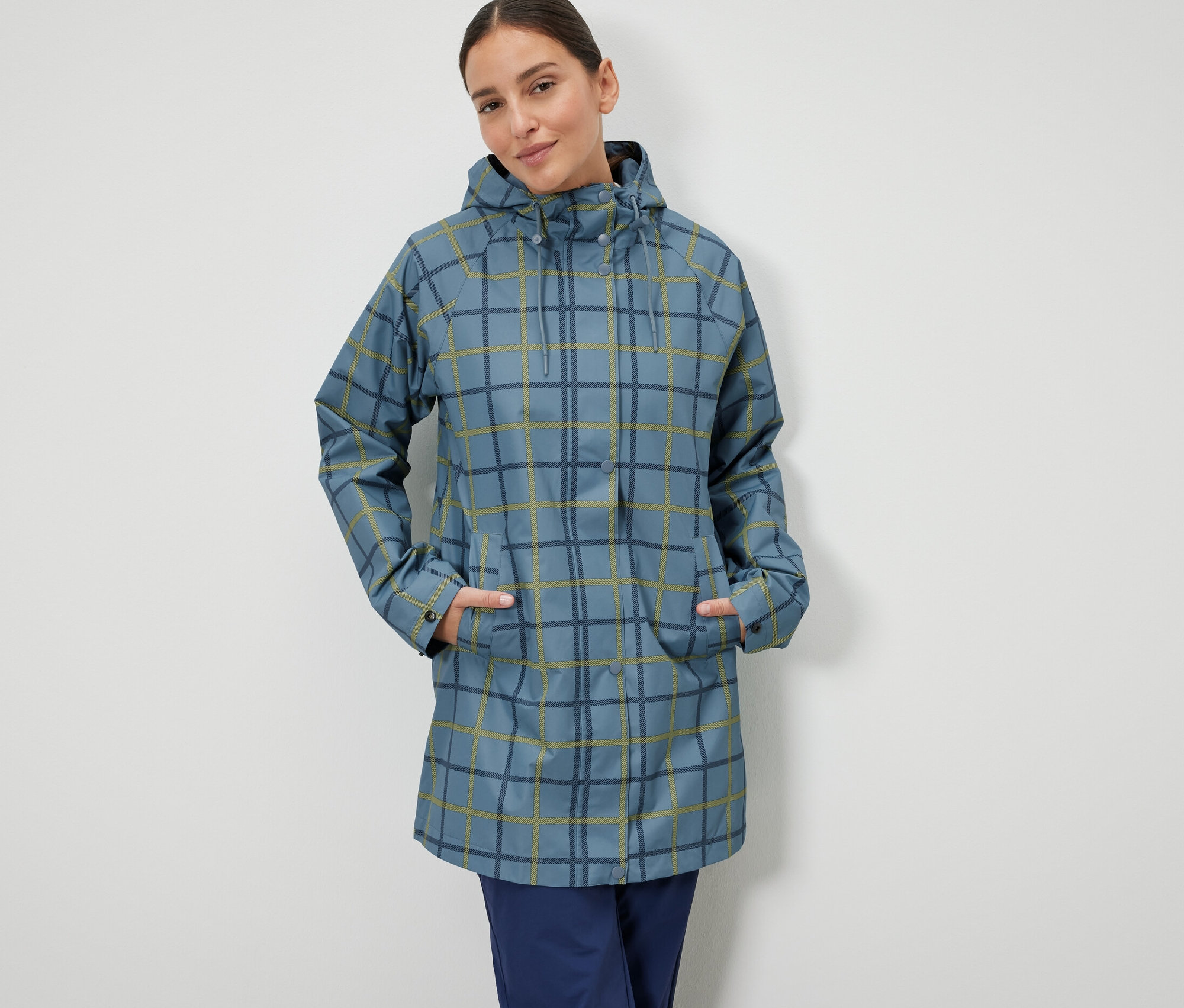 Egy nő áll, kezét a kockás mintás, kék esőkabátjának zsebébe dugva.