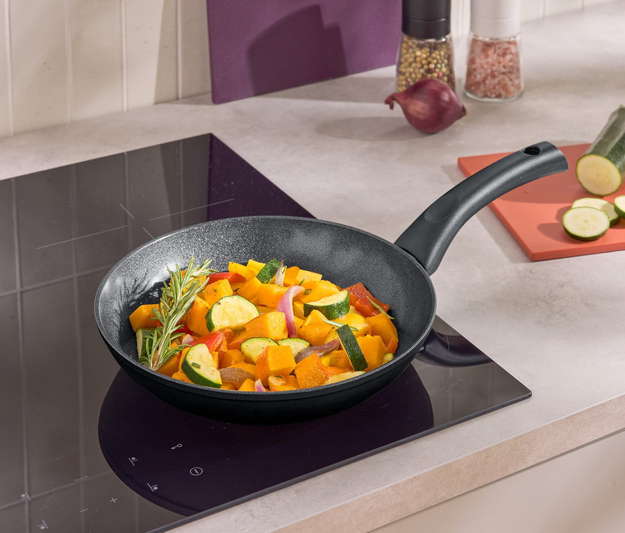 Serpenyőben sült zöldségek egy indukciós főzőlapon. A serpenyő mellett egy vágódeszkán szeletelt cukkini található.