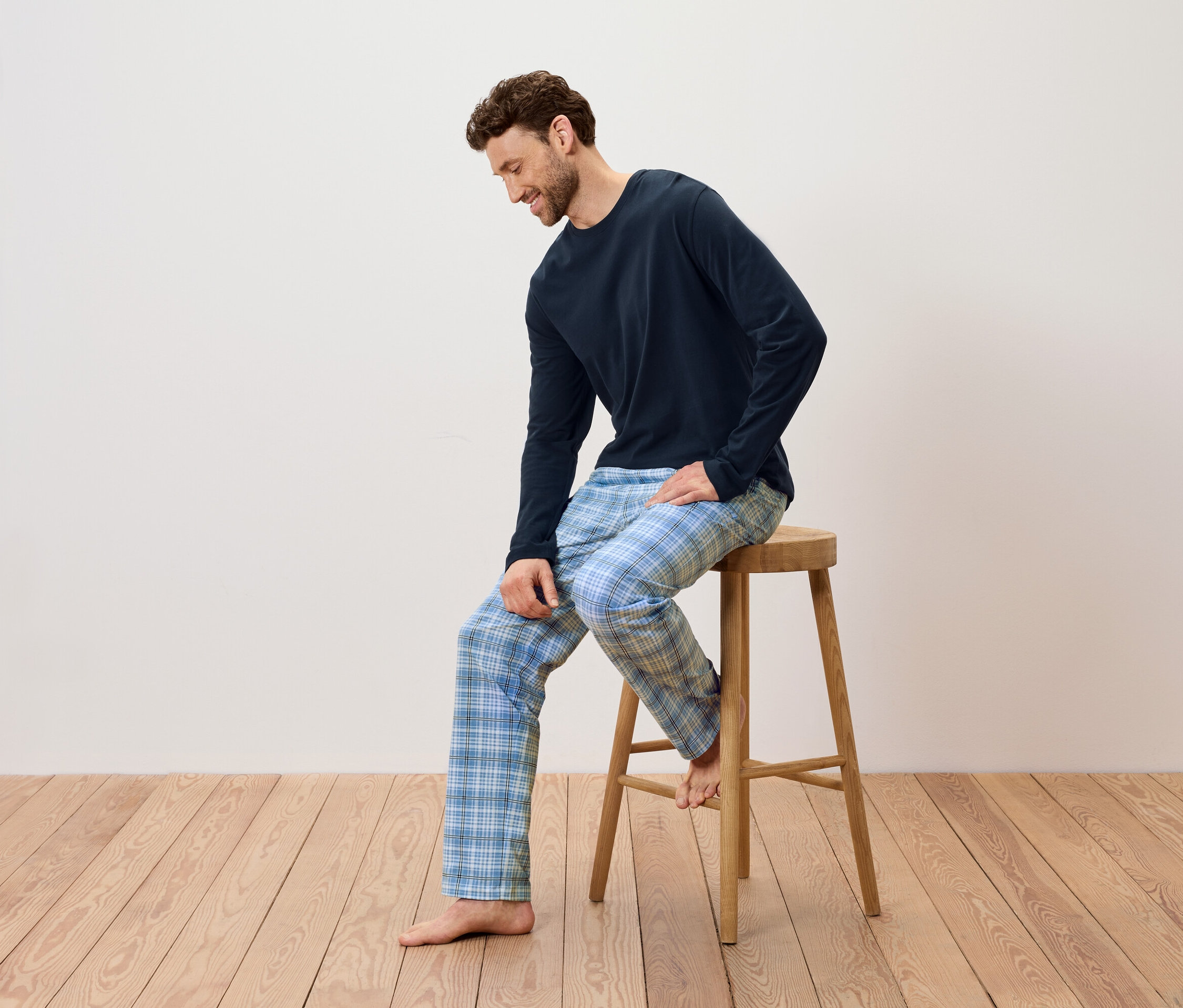 Férfi ül egy fa sámlin, sötétkék pizsamát és kék kockás nadrágot visel.