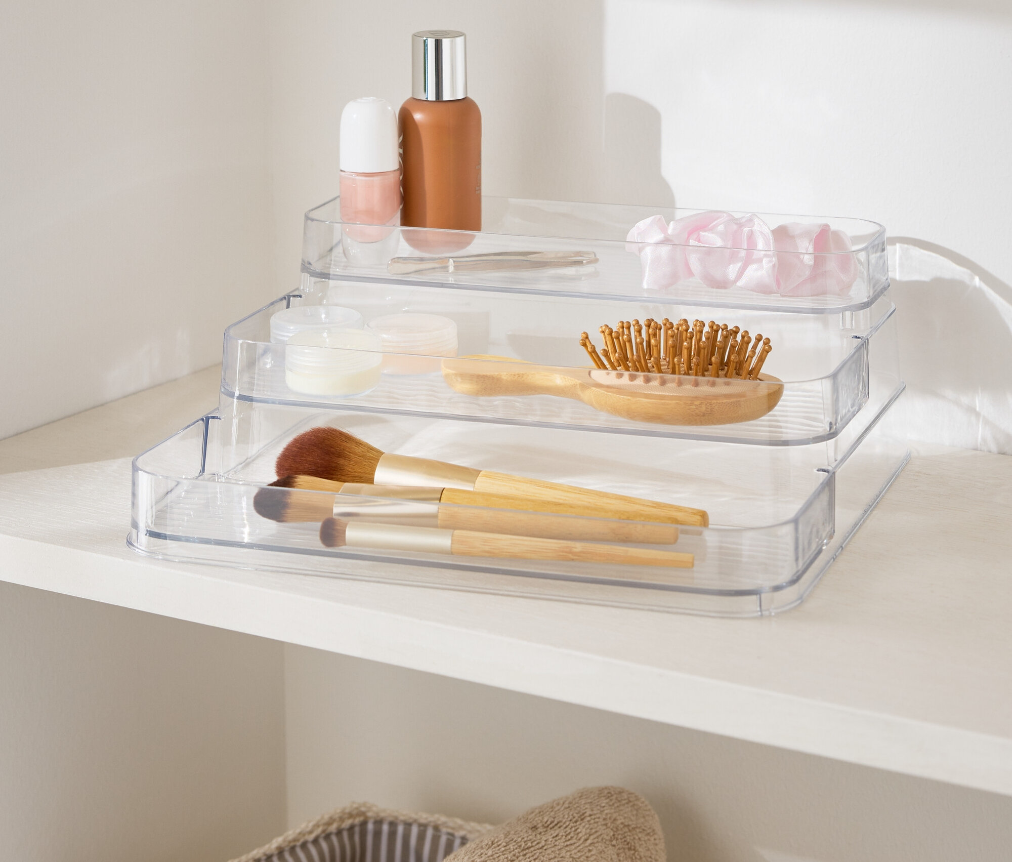 Háromszintes, átlátszó sminkrendszerező egy fehér polcon, törölközőkosarak mellett.
