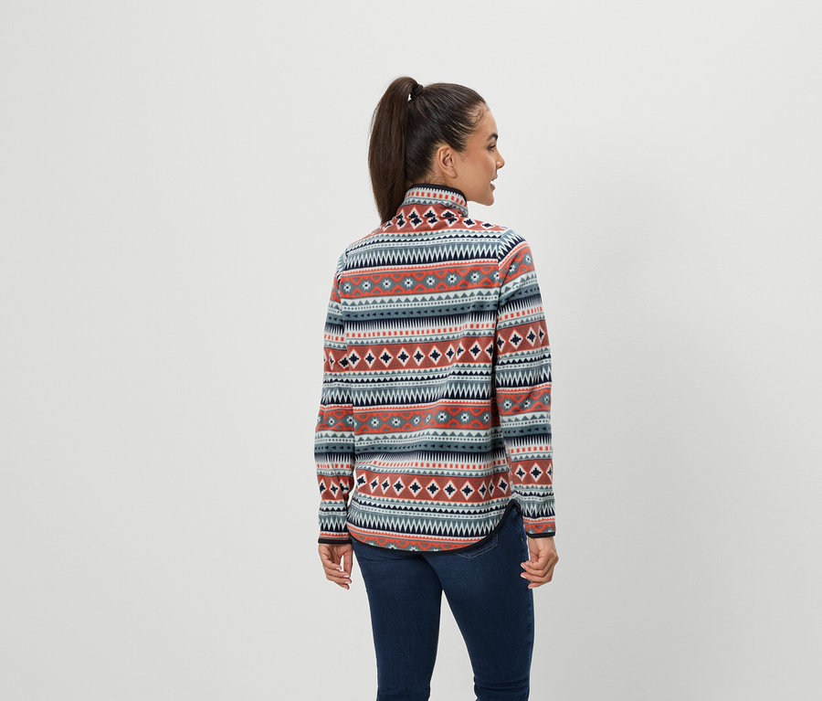 Egy barna hajú, lófarokba kötött nő hátulról áll a kamerának, kék farmernadrágot és mintás polár pulóvert visel.