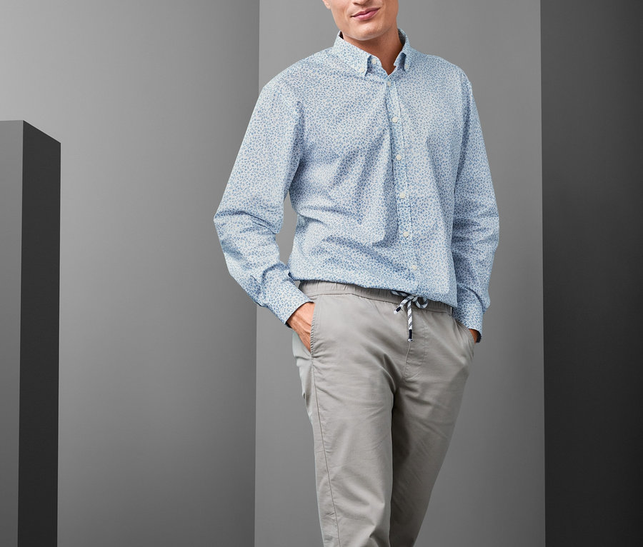 Férfi pózol, világoskék, legombolt gallérú, comfort fit férfi inget, szürke nadrágot és kék tornacipőt visel.