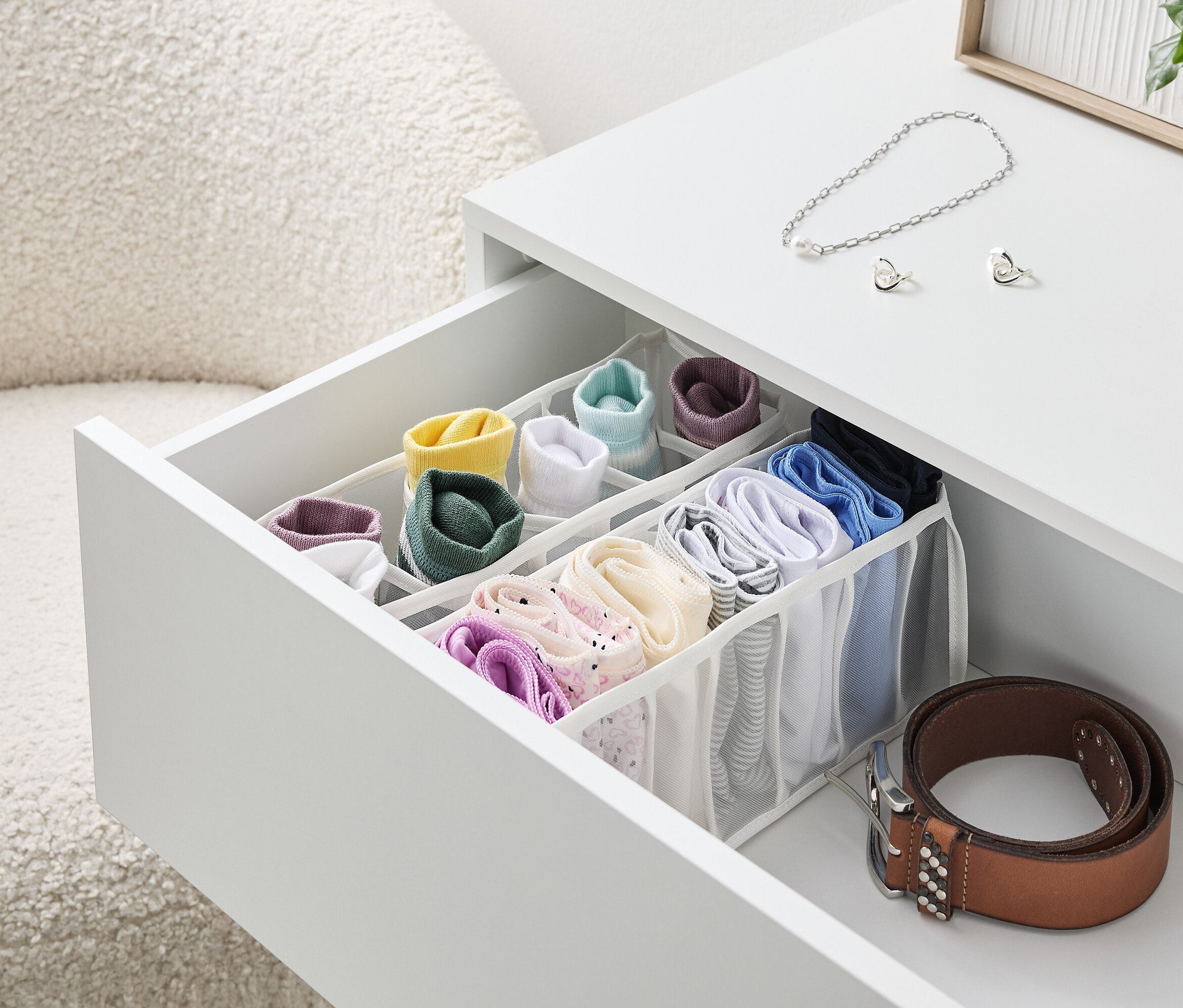 Egy nyitott fiók egy fehér asztalon, tele színes zoknikkal és fehérneműkkel egy szervezőben, mellette egy barna öv.