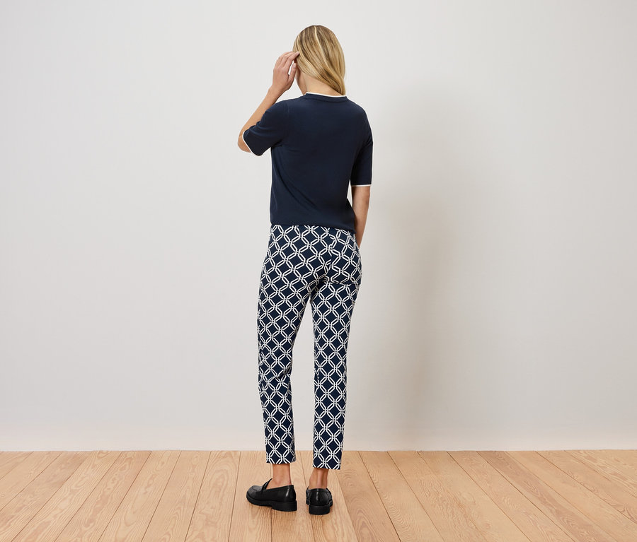 Egy nő áll háttal a kamerának, sötétkék felsőt, mintás nadrágot és fekete cipőt visel.