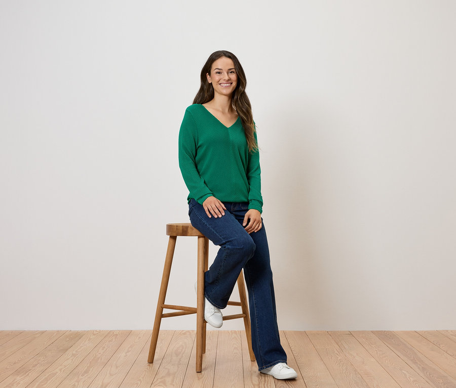 Egy nő egy fa székre ülve, zöld pulóvert és farmert visel.