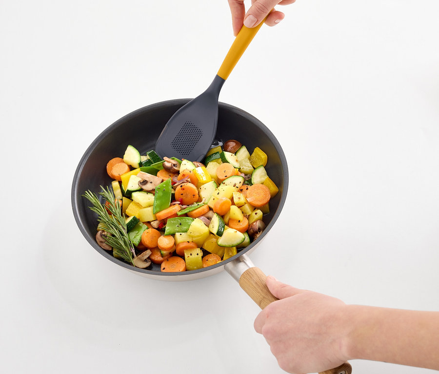 Kezek zöldségeket kevernek egy serpenyőben egy sárga, lyukacsos lapáttal.