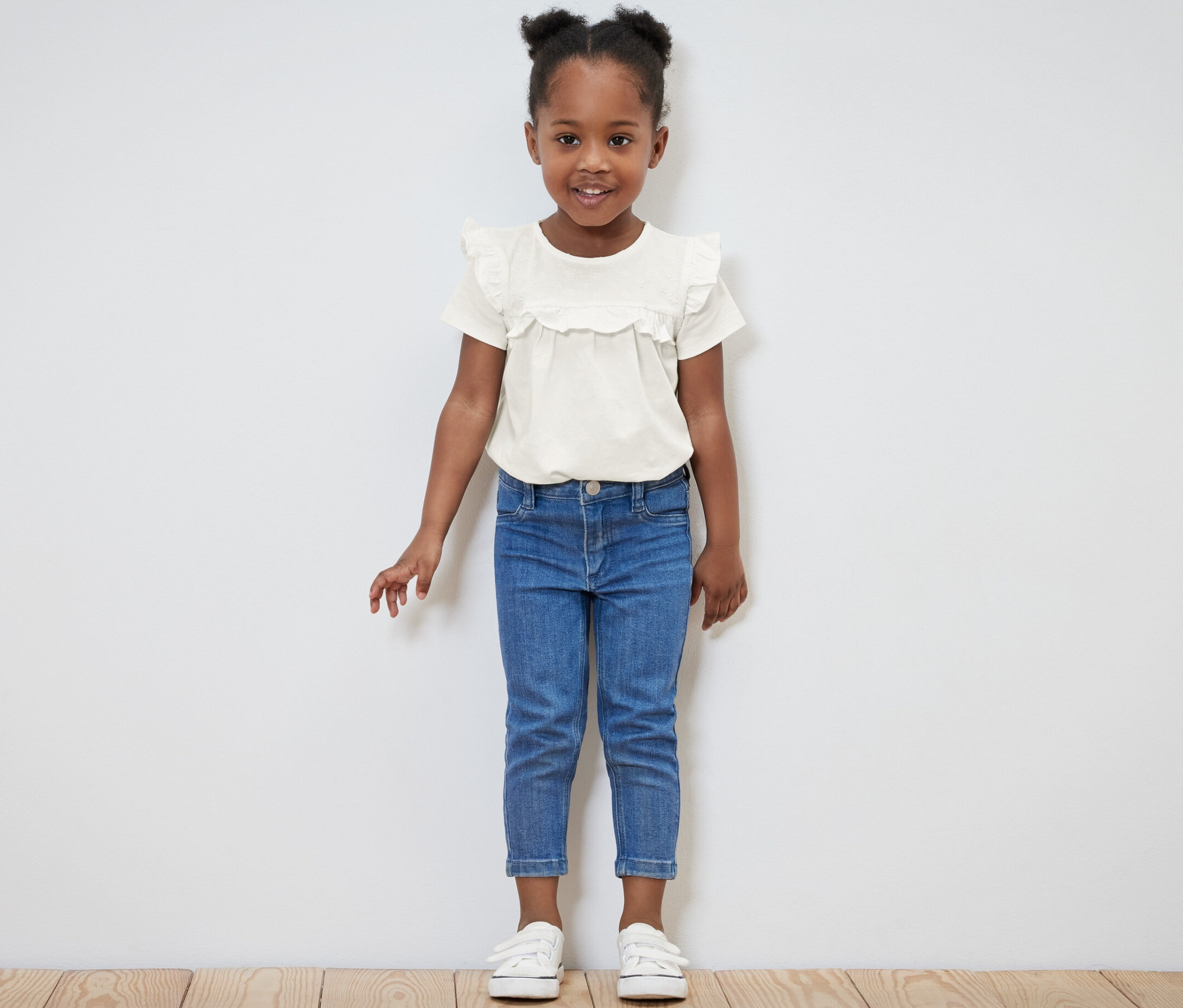 Mosolygó kislány világos gyerekpólóban és kék farmerben pózol.