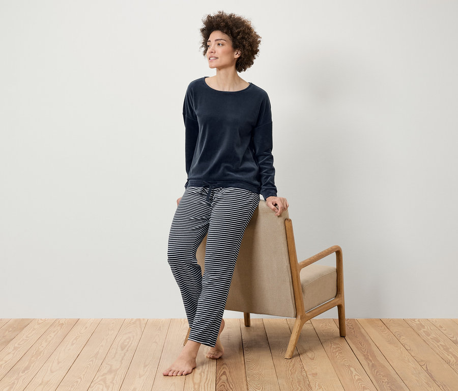 Afro frizurás nő sötétkék bársony felsőben és csíkos nadrágban pózol, egy bézs fotelnek támaszkodva a fapadlón.