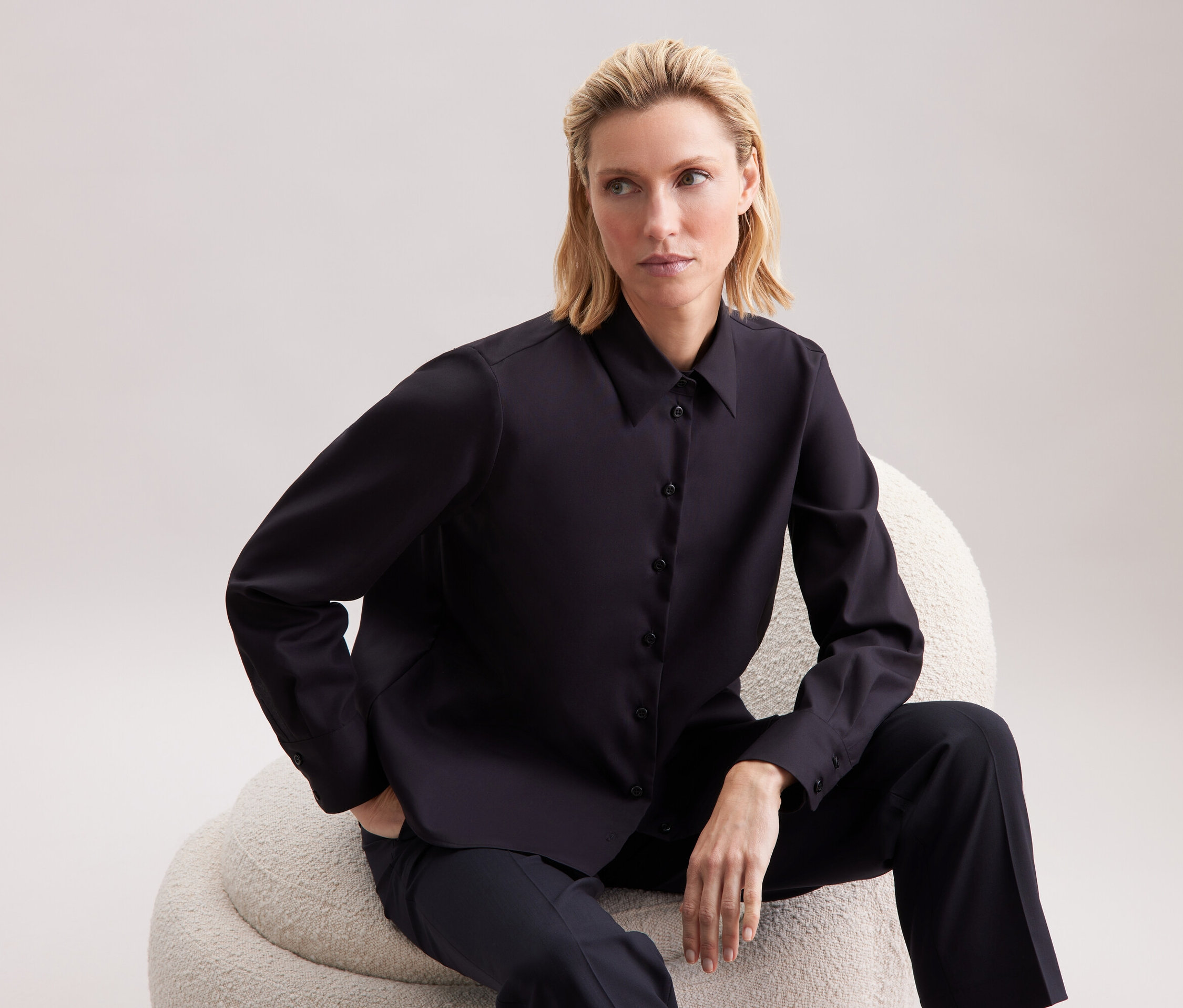 Egy szőke nő ül egy bézs széken, fekete inget és nadrágot visel.