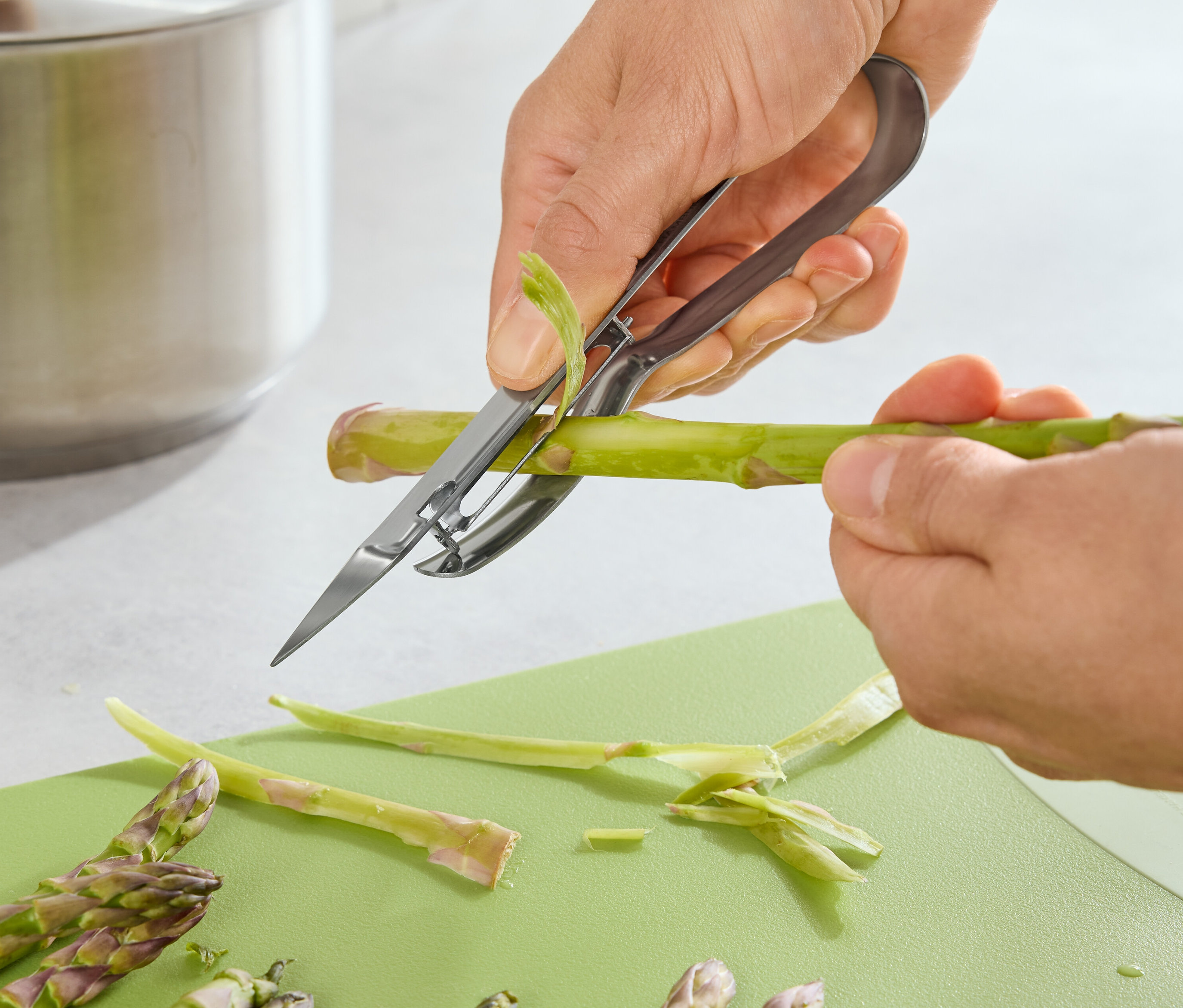 Egy személy spárgát hámoz spárgahámozóval egy zöld vágódeszkán.