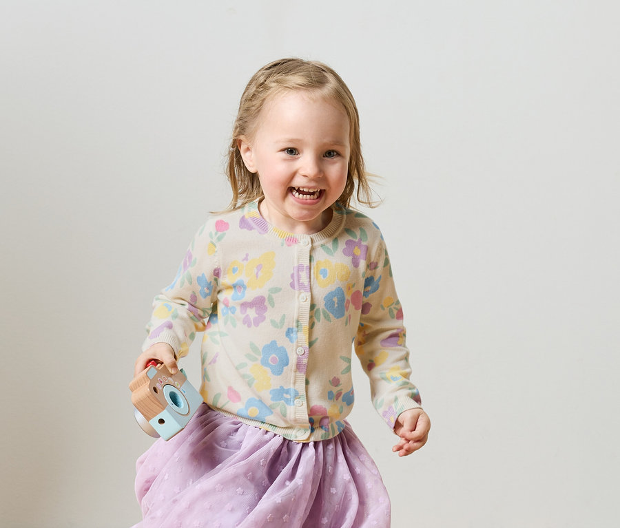 Mosolygó, fonott hajú szőke kislány virágos kardigánban és lila szoknyában játék fényképezőgépet tart.