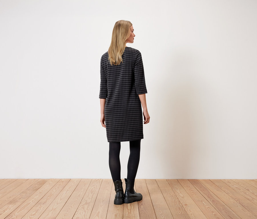 Egy nő áll háttal a kamerának, és egy houndstooth mintás ruhát, fekete leggingset és fekete csizmát visel.