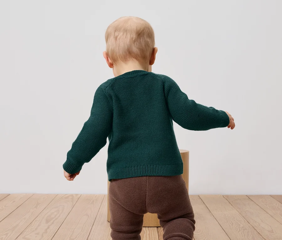 Kisgyermek zöld pulóverben és barna leggingsben áll háttal a kamerának.