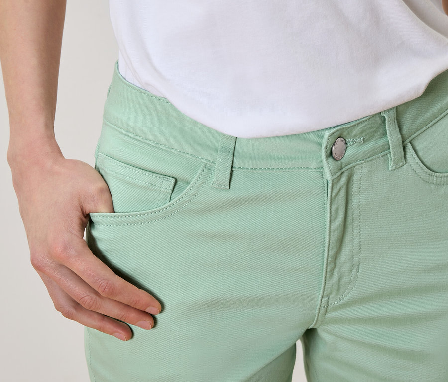 Egy személy közeli képe, akinek keze a világoszöld nadrágja zsebében van, és egy fehér pólót visel.