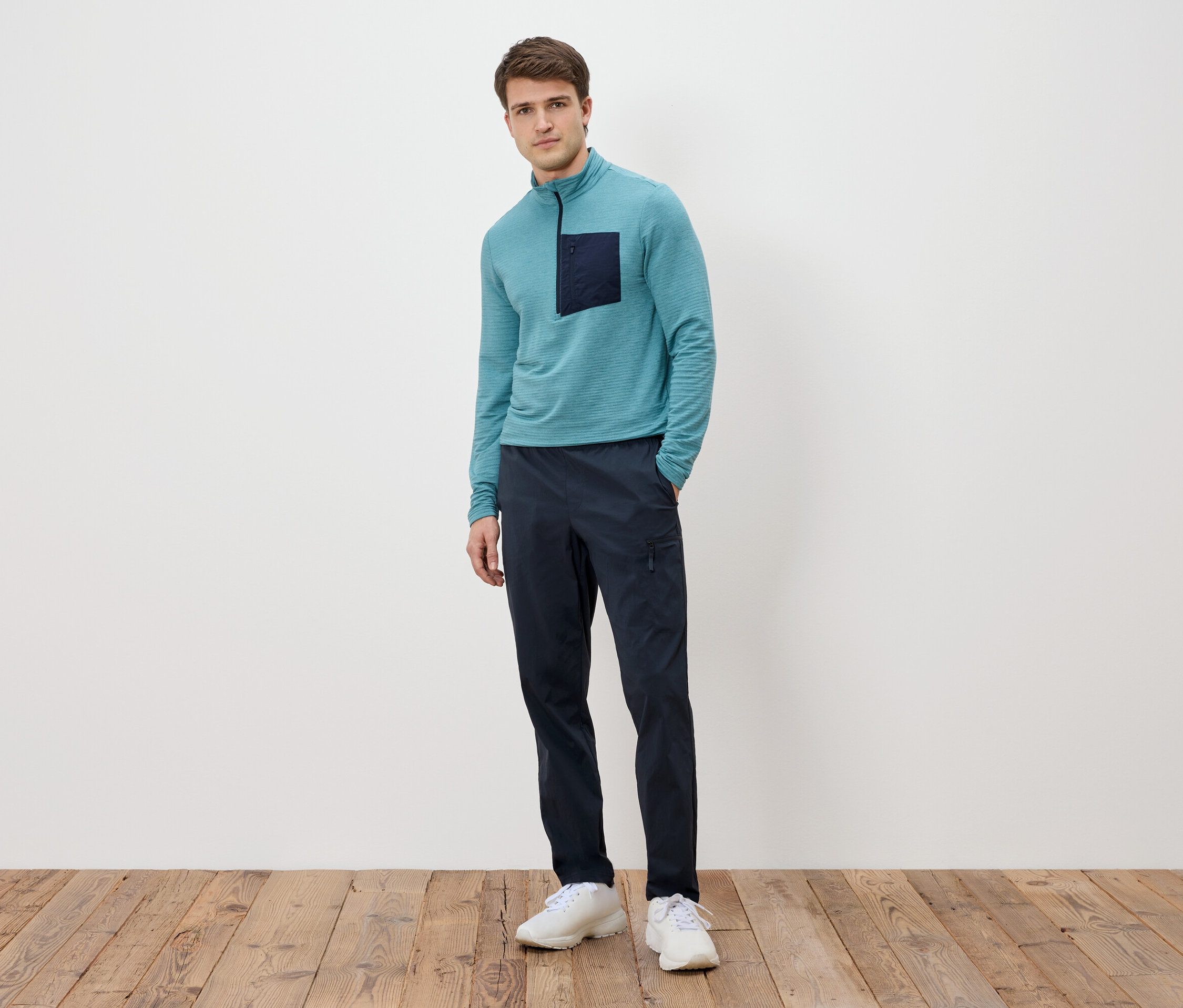 Férfi áll egy fehér fal előtt, türkizkék, hosszú ujjú pólót visel sötét zsebbel, tengerészkék nadrágot és fehér tornacipőt.