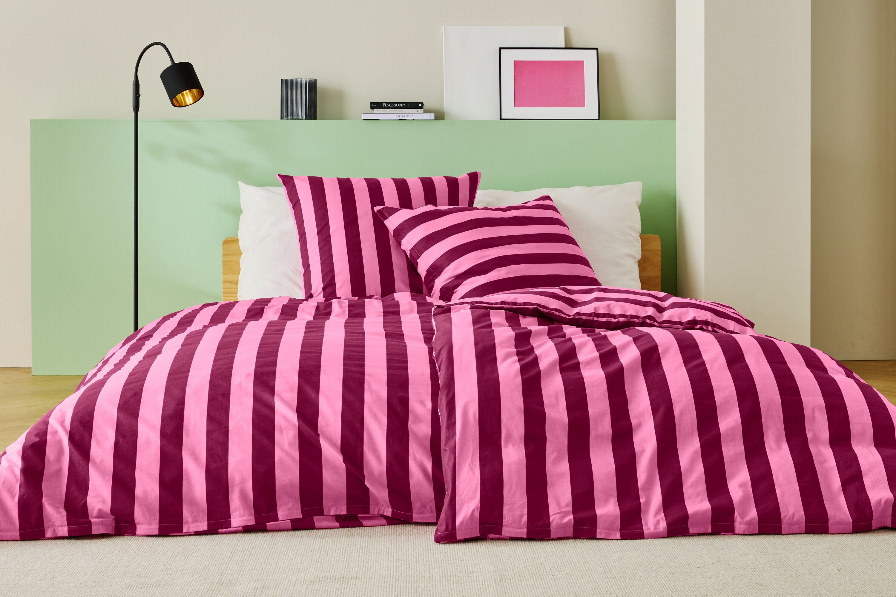 Ágy rózsaszín-bordó csíkos ágyneművel, két párnával és egy állólámpával.