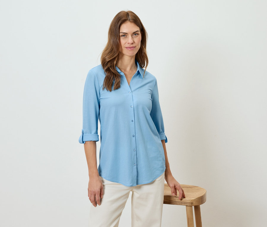 Nő világoskék inget és bézs nadrágot visel, egy zsámolyra támaszkodik.