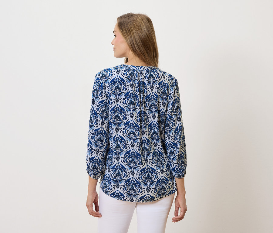 Egy hosszú, barna hajú nő kék-fehér mintás blúzt és fehér nadrágot visel.