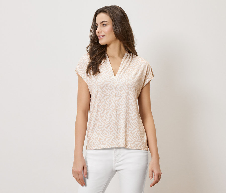 Nő visel egy világosbarna, mintás, V-nyakú felsőt és fehér nadrágot.