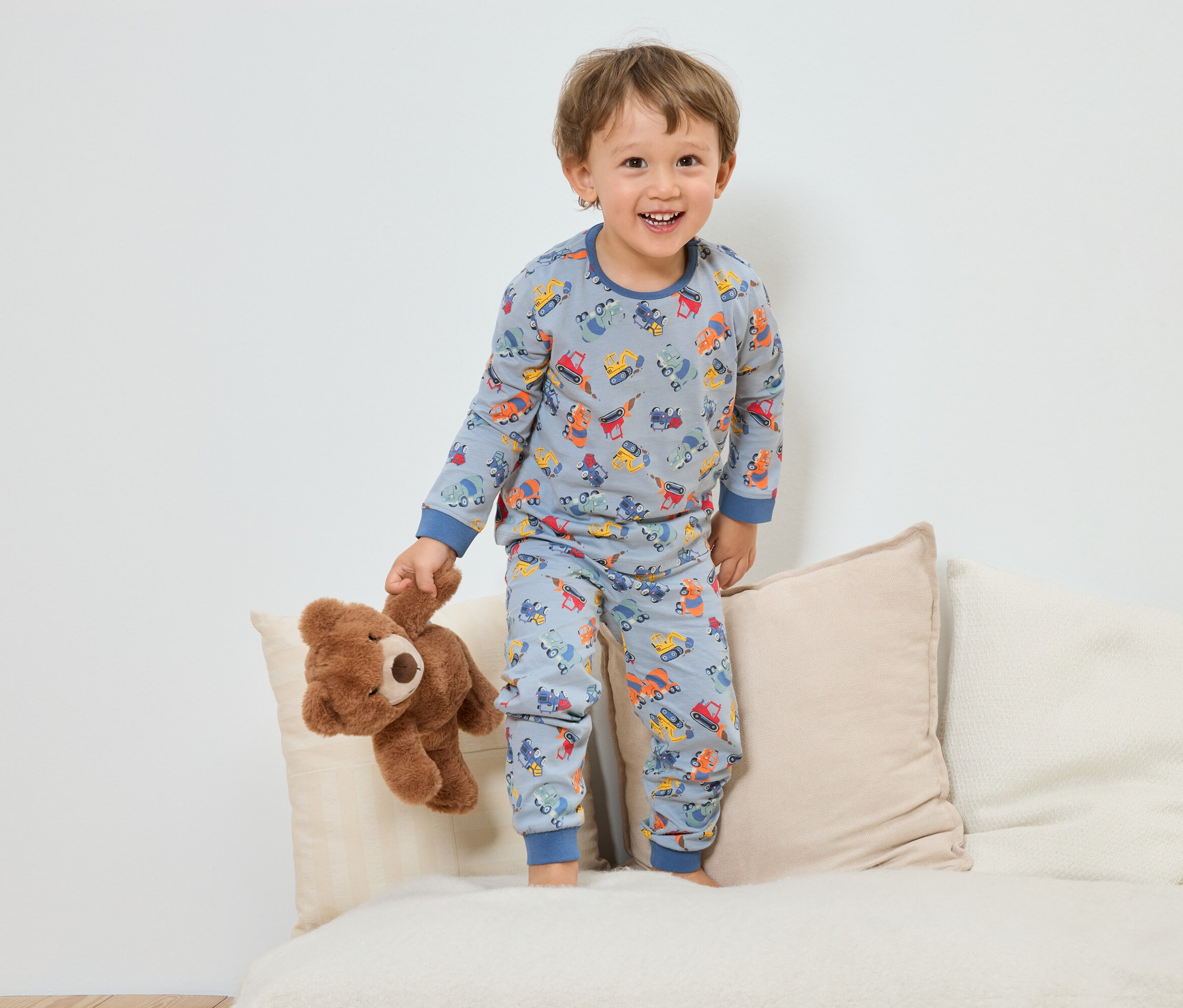 Egy mosolygó kisfiú kék, építőipari jármű mintás pizsamában áll egy plüssmackóval egy kanapén.