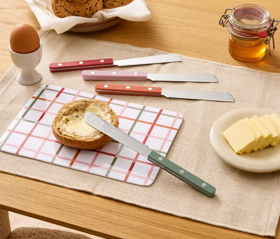 Egy asztalon megkent zsemle, négy kés, vaj, méz, tojás és kenyér található.