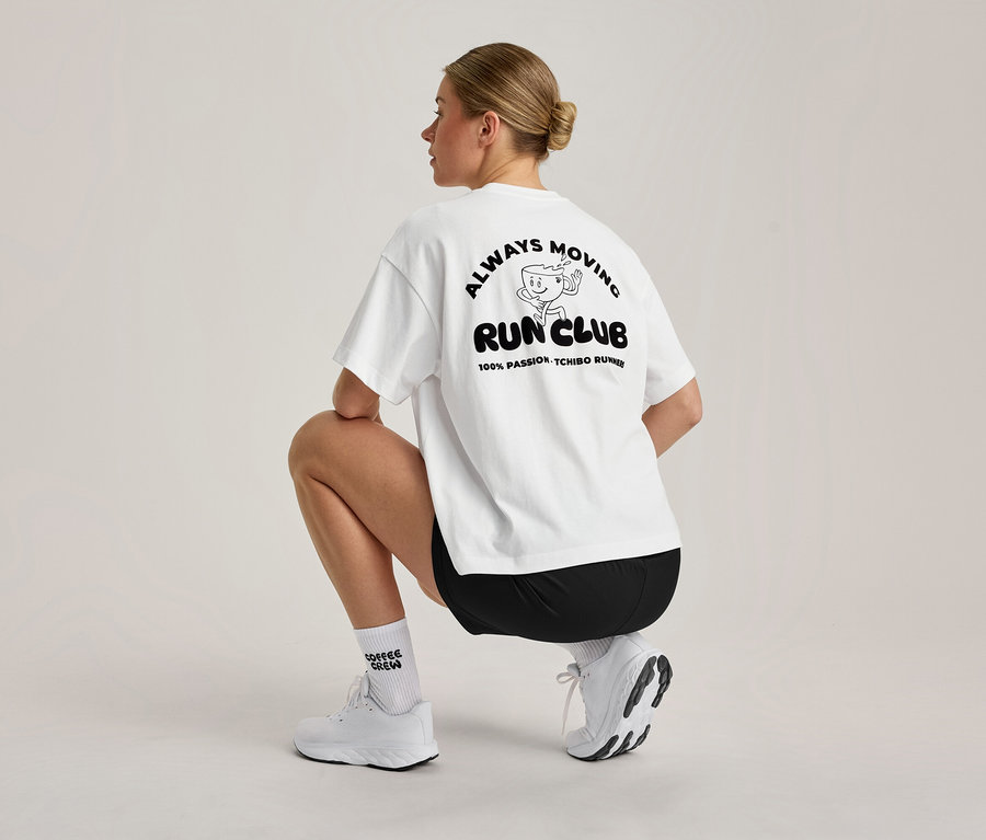 Egy szőke kontyos nő guggol, hátulról nézve. Fehér, oversized sportfelsőt visel fekete 'ALWAYS MOVING RUN CLUB' grafikával a hátán, fekete rövidnadrágot, fehér zoknit és fehér sportcipőt.