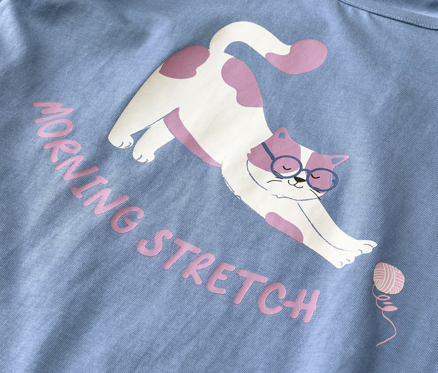 Világoskék anyag egy fehér, rózsaszín foltos, kék szemüveges macska grafikájával, amint nyújtózkodik. Rózsaszín szöveg olvasható: "MORNING STRETCH" egy kis rózsaszín fonalgombolyag mellett.
