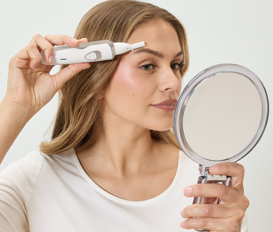 Egy nő fehér epilátorral szedi a szemöldökét, miközben egy kerek tükörbe néz.