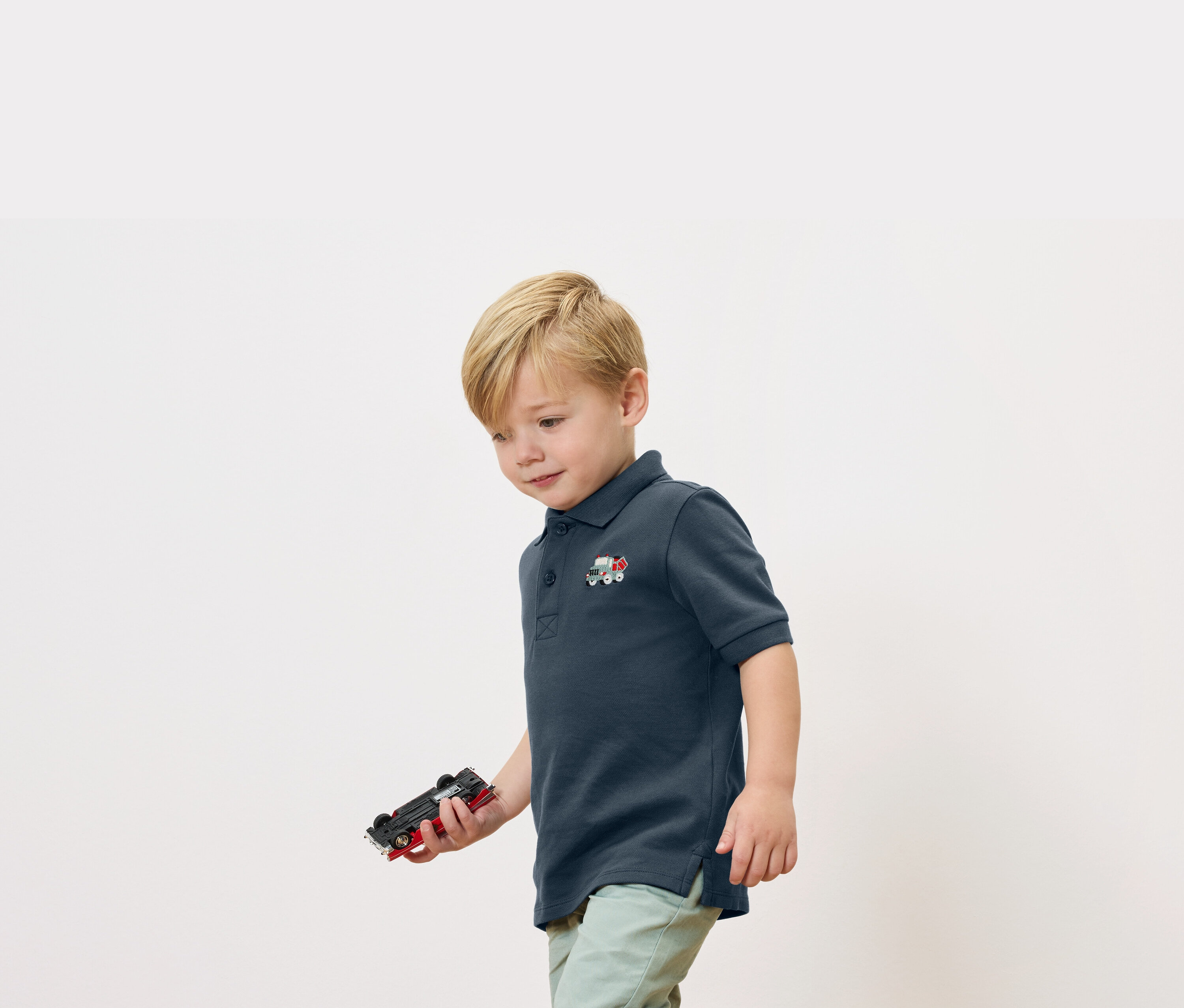 Egy szőke fiú játékautót tart a kezében, és egy sötétkék pólót visel egy kis vonatmintával.