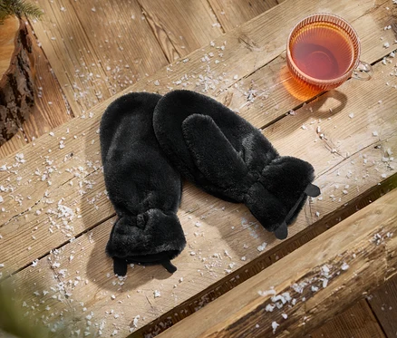 Egy pár fekete műszőrme kesztyű egy csésze tea mellett, havas fafelületen, fenyőtobozzal és ággal.