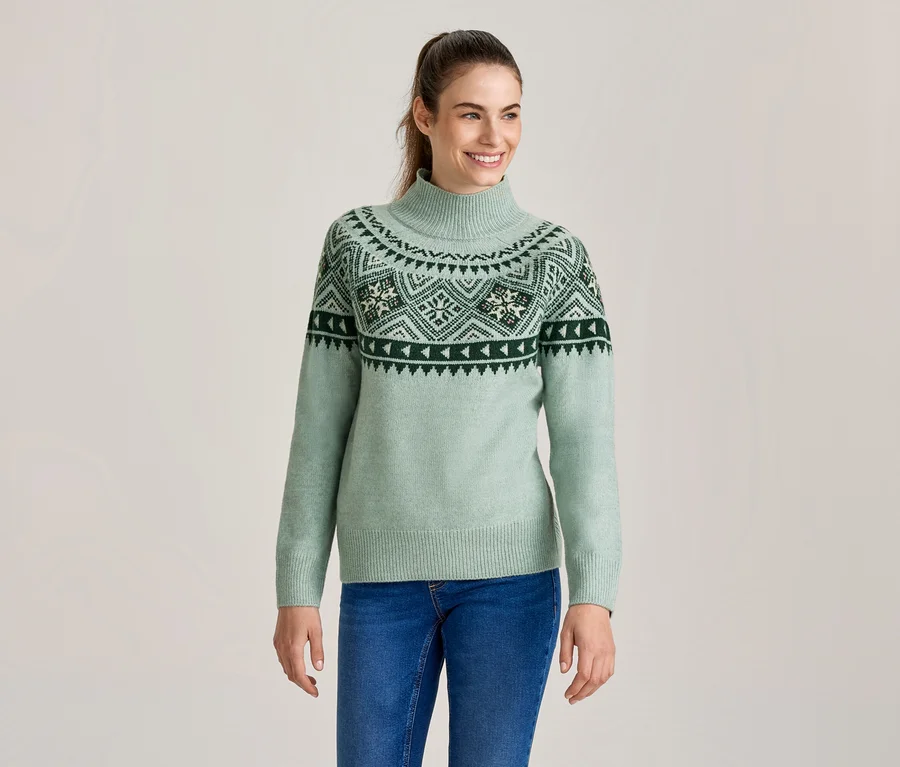 Mosolygó nő világoszöld garbó pulóverben, sötétzöld és fehér Fair Isle mintával, kék farmernadrággal.