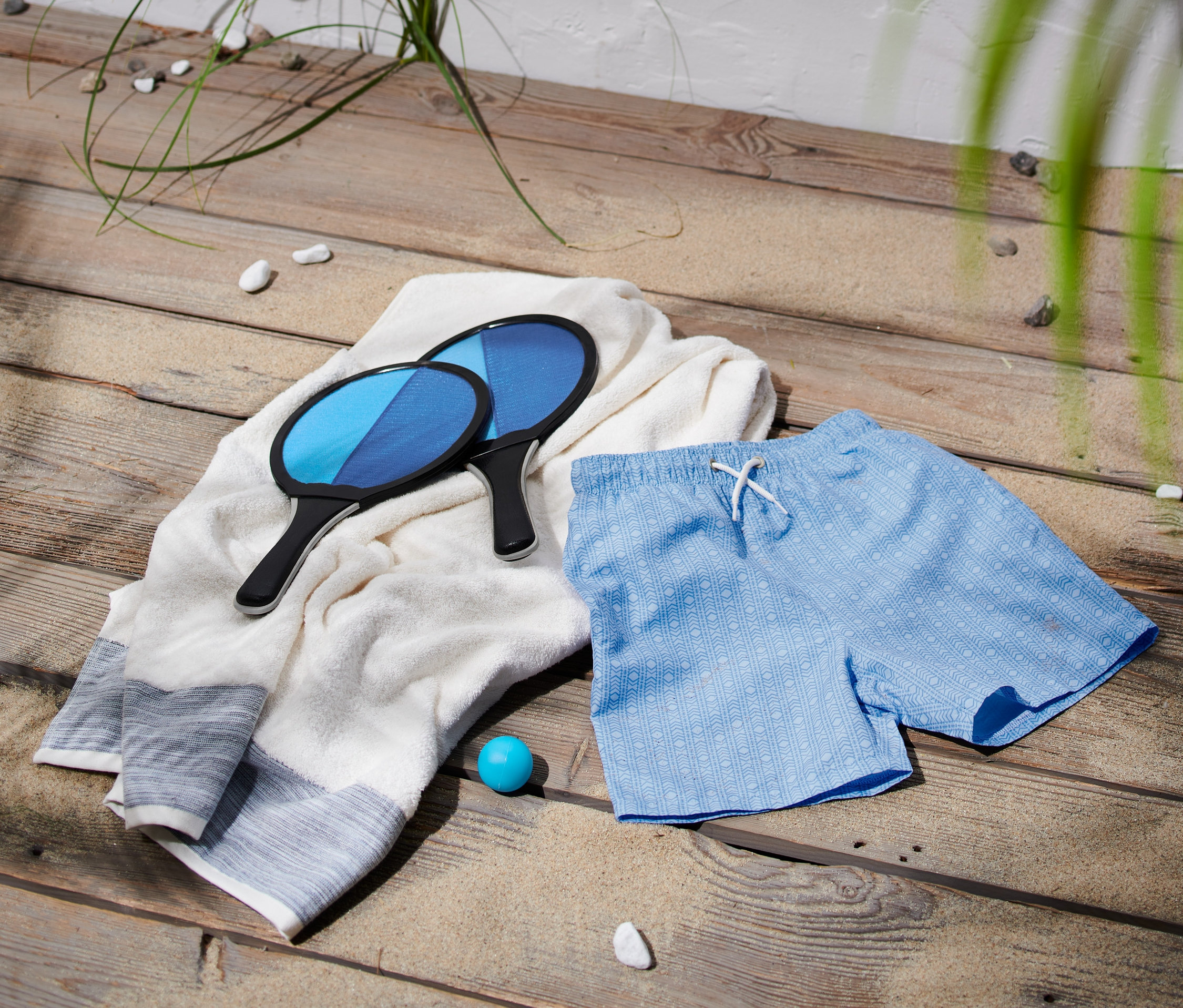 Két strandütő fekszik egy fehér törölközőn egy kék, mintás fiú fürdőnadrág mellett.