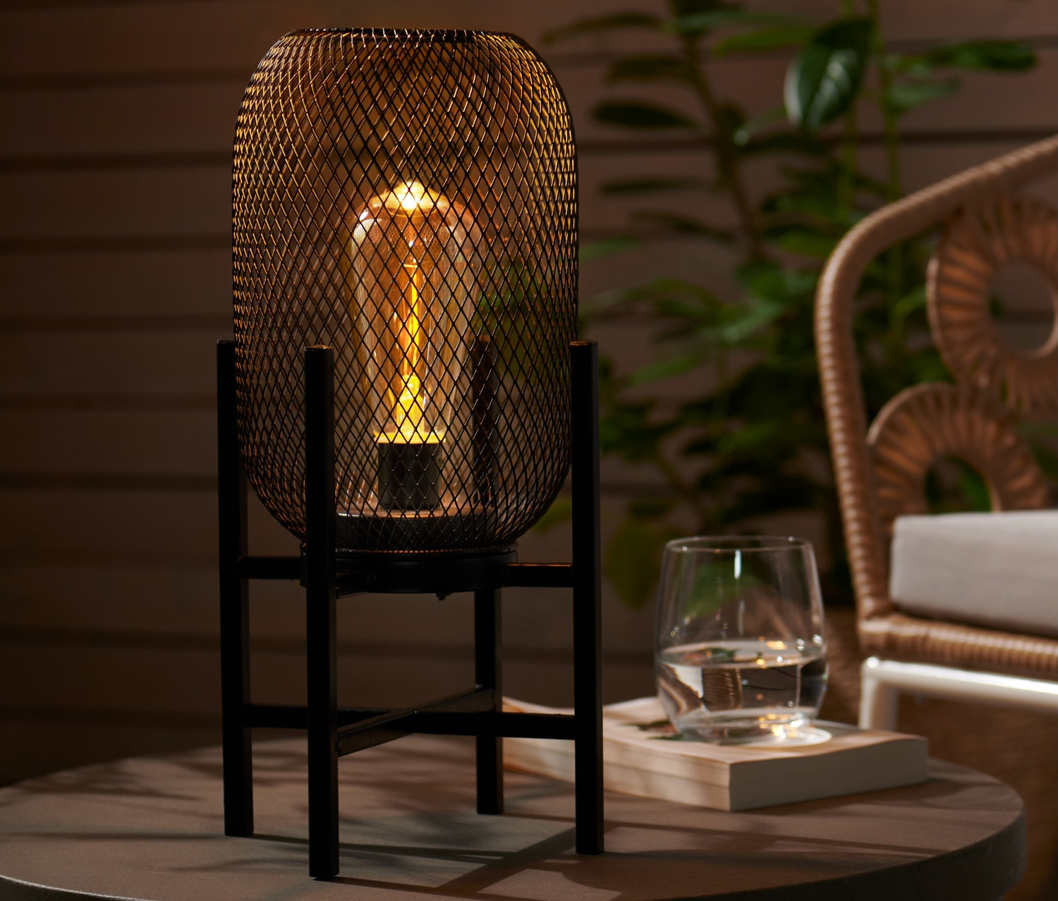 Egy kültéri LED-es asztali lámpa áll egy kerek asztalon egy pohár víz mellett. A háttérben egy rattan fotel látható.