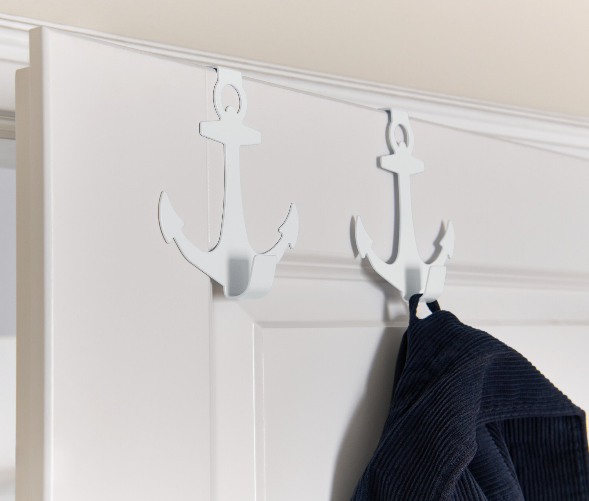 Két horgony alakú fehér ajtófogas lóg egy fehér ajtón, az egyiken kék ruha lóg.