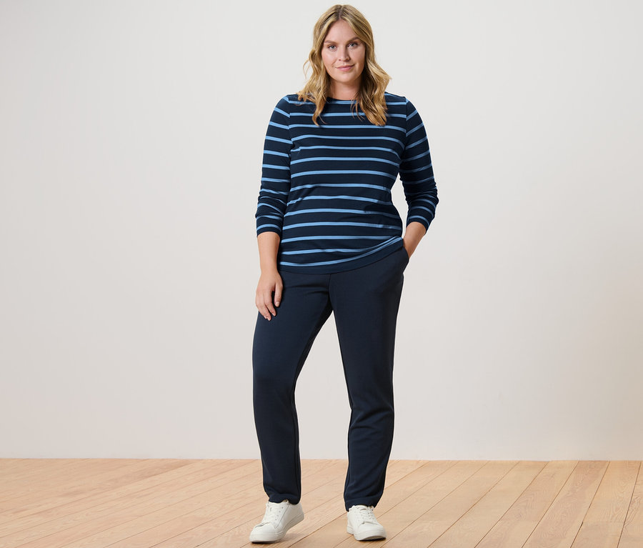 Nő áll sötétkék női lezser nadrágban és csíkos, sötétkék/világoskék női hosszú ujjú felsőben.