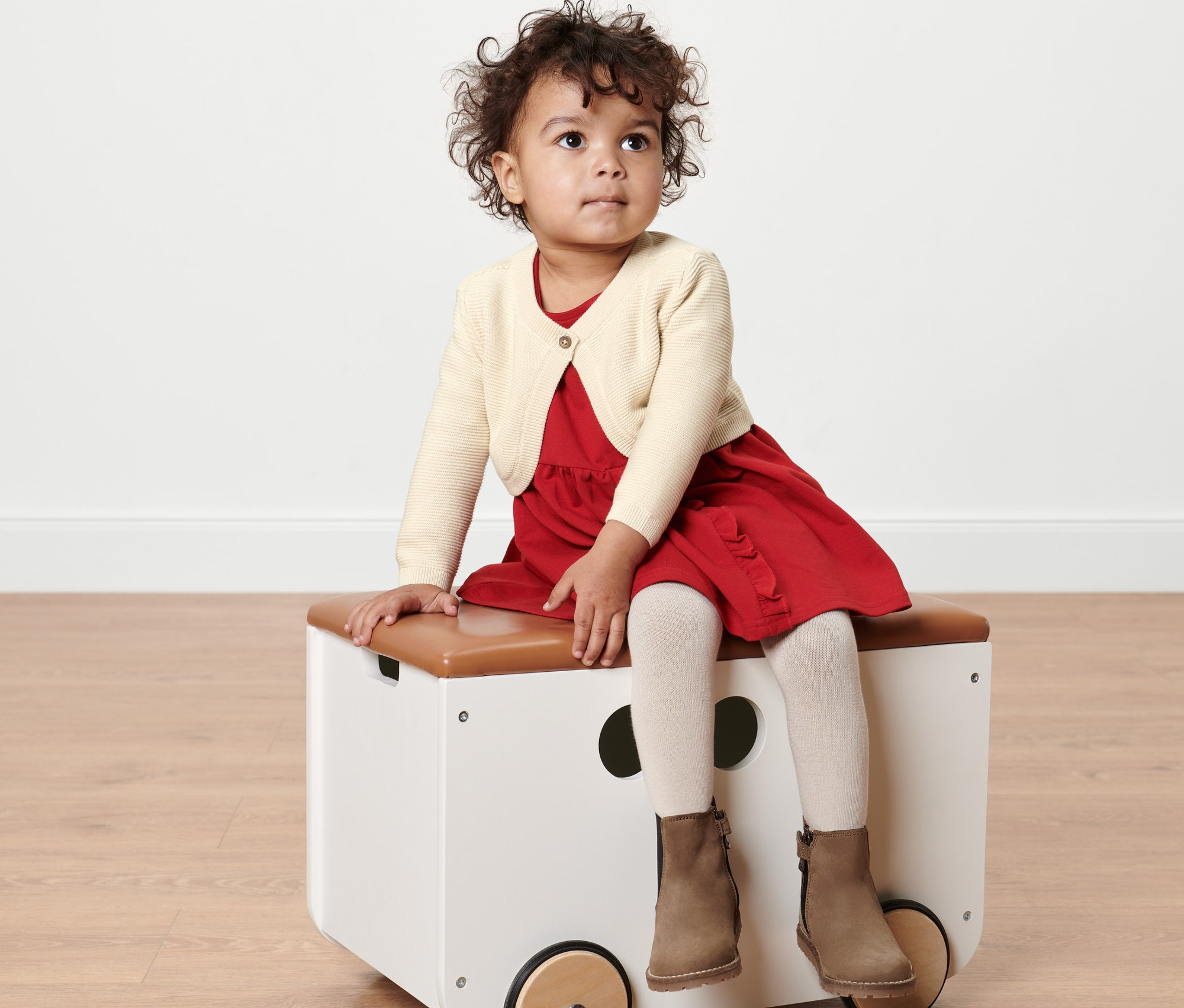 Kislány ül a játéktárolón, krémszínű kötött boleró van rajta.