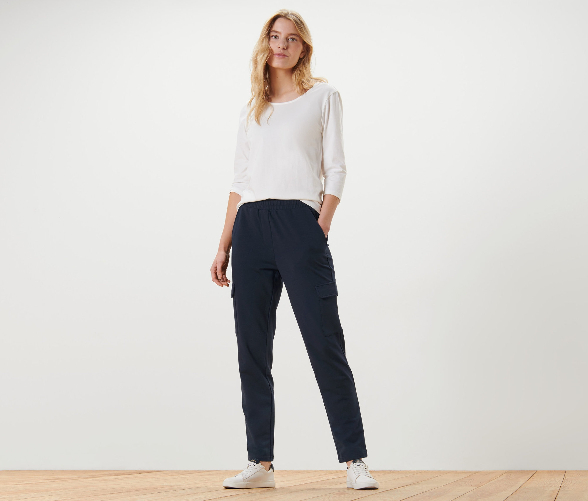 Egy nő áll, kezét a sötétkék oldalzsebes lezser nadrág zsebébe dugva.